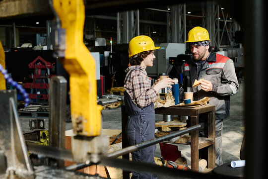Two industrial workers wearing safety helmets and holding coffee cups, discussing during a break