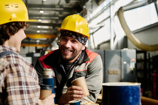 Two factory workers in protective gear are smiling while sharing a coffee break in an industrial setting, revealing camaraderie and teamwork