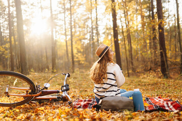 A young woman sits on a blanket surrounded by colorful autumn leaves, smiling happily as she holds a warm drink, with a bicycle nearby in a serene forest.