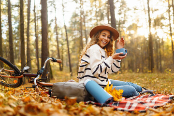 A young woman sits on a blanket surrounded by colorful autumn leaves, smiling happily as she holds a warm drink, with a bicycle nearby in a serene forest.