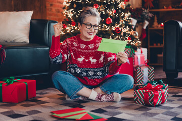 Full size body photo of funny retired woman in deers print jumper sitting on carpet and reading mail at christmas atmosphere indoors