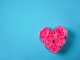 A heart-shaped container filled with pink roses sits on a blue background. The roses are arranged in a way that they look like they are blooming, and the container itself is clear