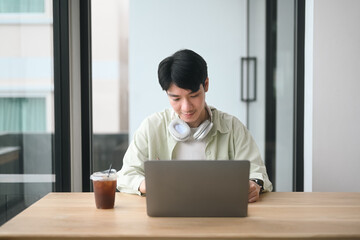 A Young Asian man working intently on a laptop in a coffee shop, with headphones and an iced coffee nearby, Focused Work at a Coffee Shop