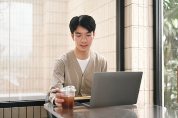 A man is working intently on his laptop while holding a cup of iced coffee, embodying the modern freelance or remote work lifestyle