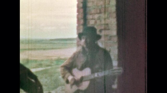 Male cowboy playing guitar near window on natural landscape background. Old outdated western style. Man singing song. Artist guitarist in hat play music instrument. Vintage color film. Retro archive