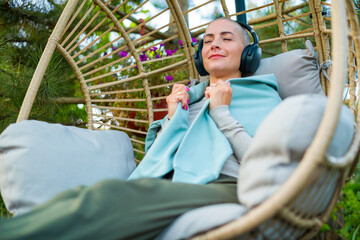 Young female cancer patient relaxing in hanging chair on the garden patio, listening to music....