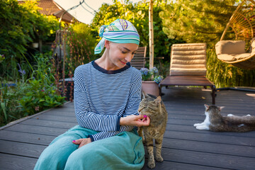 Young female cancer patient wearing headscarf petting her cat sitting by her side on the garden...