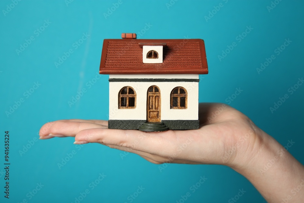 Wall mural Woman with house model on light blue background, closeup