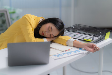 A young female employee of Asian descent, seated in her office, expresses weariness from the...