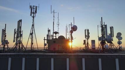 Silhouette of electromagnetic towers with satellite dish, microwaves and panel antennae at rooftop station during golden hour