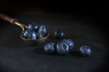 Vintage brass spoon with blueberries on a black slate plate, dark and moody still life with fruits,...