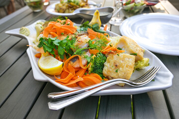 White platter in fish shape with various fried fish fillets and vegetables on a large dining table with dishes and plates in an outdoor restaurant at the sea, selected focus