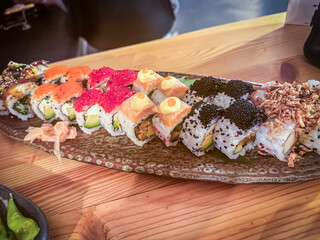 A variety of sushi rolls arranged on a long ceramic platter on a wooden table.