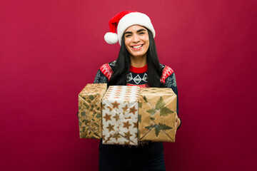 Happy young woman wearing a christmas sweater and santa hat holding presents on a red background