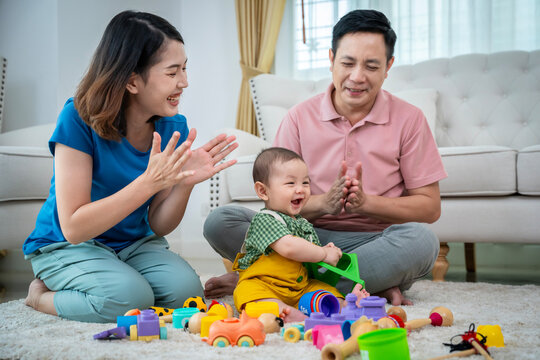 A family of three, a man and two women, are playing with toys on the floor. The baby is holding a toy truck and the parents are clapping. Scene is happy and playful