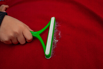 A hand is using a green and white pet hair remover tool to clean hair off a red fabric surface.
