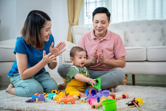 A family of three, a man and two women, are playing with toys on the floor. The baby is holding a toy truck and the parents are clapping. Scene is happy and playful