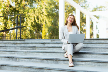 Stylish professional woman working on her laptop while seated on modern outdoor steps in a bustling urban setting during the golden hour