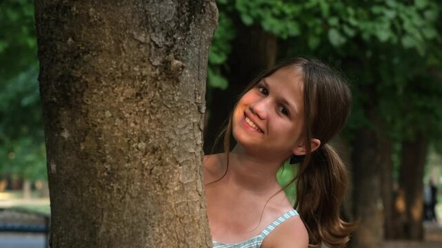 Smiling girl 11 years old peeks out from behind a tree in the park. Happy child playing. Childhood and dreams concept