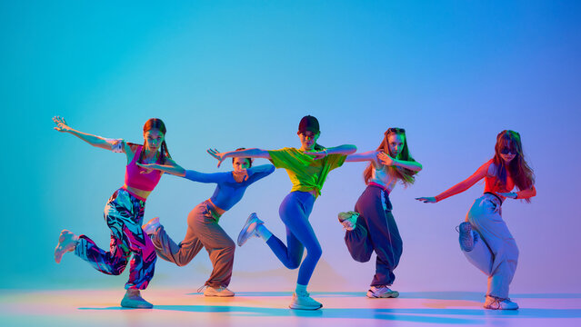 Dressed stylish, sport cloths teenage girls performing hip-hop in neon light against gradient studio background. Synchrony. Concept of sport and hobby, childhood, friendship, motion. Dance lessons.