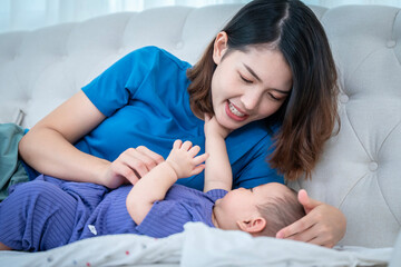 A woman is sitting on a sofa with her baby on her lap. The baby is wearing a purple dress and is drinking milk from a bottle.