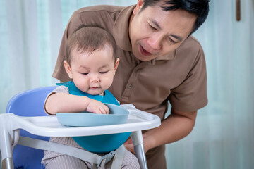 A man is feeding a baby in a high chair. The baby is wearing a bib and is eating from a bowl. The...