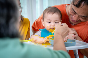 A woman feeds a baby in a high chair. The baby is wearing a bib and is eating food. The scene is...