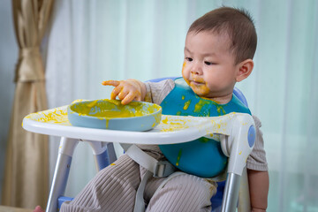 A baby is sitting in a high chair with a yellow mess on his face. The baby is wearing a green bib...