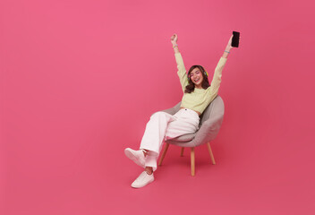 Cheerful young asian teen woman in wireless headphones sitting in armchair, listening to music, raising her arm, dancing to favorite song, enjoying popular soundtrack isolated on pink background.