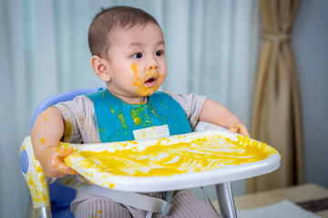 A baby is sitting in a high chair with a yellow mess on his face. The baby is wearing a green bib...