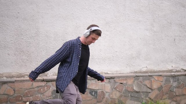 young man in blue checkered shirt and black t-shirt listens to music with large white headphones and dance. He stands outdoors against light-colored textured wall with green leafy plants background