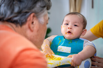 A baby is sitting in a high chair with a yellow mess on his face. The baby is wearing a green bib...