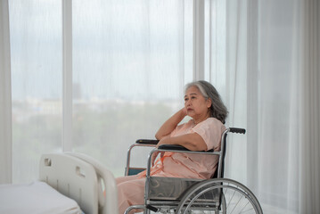 Depressed senior Asian woman patient in hospital sitting in wheelchair by the window, upset disabled patient thinking and looking out window, feeling lonely, nostalgia and melancholy