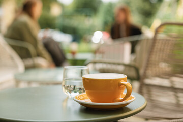 A ceramic cup with a hot drink and a glass of water on the table on the summer terrace with unrecognizable girls sitting at the table in the background.