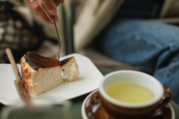 Close-up of a young woman enjoying a slice of Basque cheesecake and a cup of buckwheat tea on a summer terrace.
