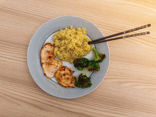 A top view of a meal with grilled chicken, quinoa, and roasted broccoli, served with chopsticks on a dotted plate over a wooden table.