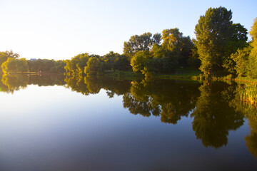 Evening in the Loshitsky Park in Minsk. The trees are reflected in the calm water of the lake. The landscape in the evening light of the Sun.
