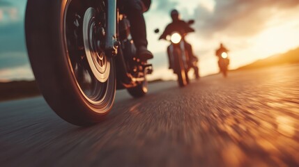 Three motorcycles drive on a road at sunset, captured from a low angle focusing on the front wheels, with the sun setting in the background.