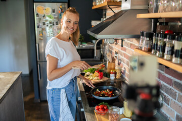 Smiling young woman cooking and filming with smartphone in modern kitchen