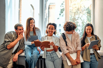 Five smiling diverse confident smart college students standing near window reading books and...