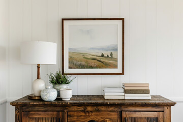 A large framed landscape painting hanging on the wall of a farmhouse, above a rustic wood sideboard with books and white lamps, in neutral tones with pops of greenery, farmhouse-style home decor photo