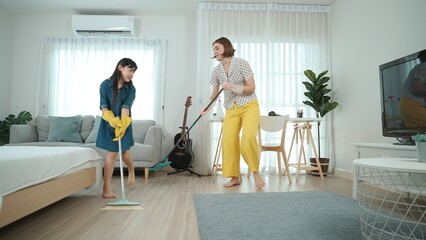 Caucasian mom and asian child doing housework together and dancing to music. Attractive mother and daughter cleaning modern bedroom while sing along and jump to relaxing music. Lifestyle. Pedagogy.