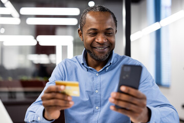 Smiling man in office holding credit card and using smartphone for online shopping. Bright, modern setting creates professional and approachable atmosphere.