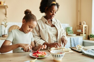 Medium shot of African American mother and little daughter cooking together cutting strawberries and bananas for healthy breakfast at cozy beige kitchen