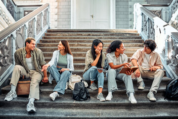 Fototapeta premium Multicultural students sitting together on the stairs and talking. They hold books, notebooks, backpacks and laptops.