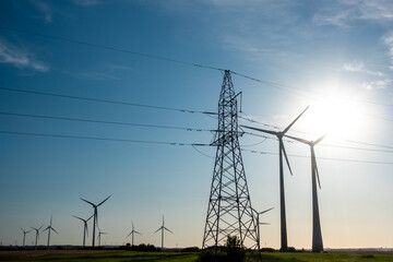 Wind turbines and high voltage electric pylon against blue sky. Sun shining behind the turbines