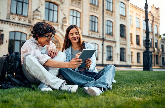Two young students are sitting on the grass on the campus talking and preparing for classes. Real student life.