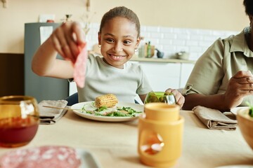 Medium shot of smiling African American girl eating roasted corn at table treating herself to slice of sausage during family dinner at beige kitchen