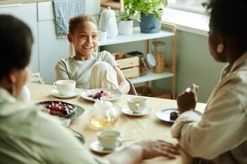 Young smiling Black girl sitting at table talking to family over cup of tea with delicious dessert on little plate at sunlit kitchen, copy space
