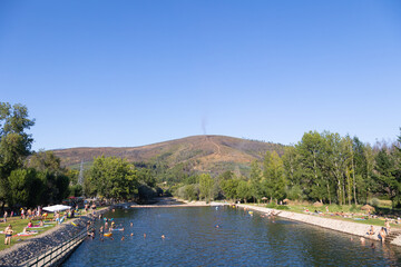 Obraz premium A scenic river beach at Praia Fluvial de Valhelhas in Portugal, with families enjoying swimming and sunbathing under a clear blue sky. This serene summer destination offers a perfect mix of relaxation